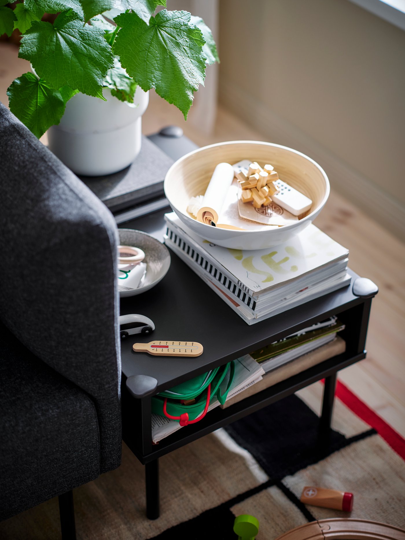 A TUNSTA side table, with books and a plant on it and with UNDVIKA corner bumpers on its corners, stands on a RASTPLATS rug.