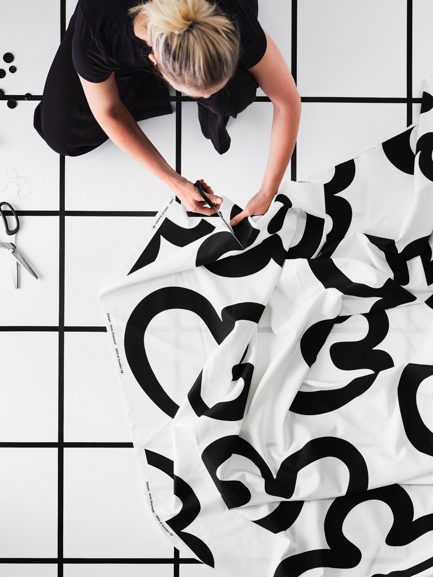 En person sitter på golvet, håller i en stor bit förskuren KLIPPBRÄCKA tyg och klipper den med en sax.