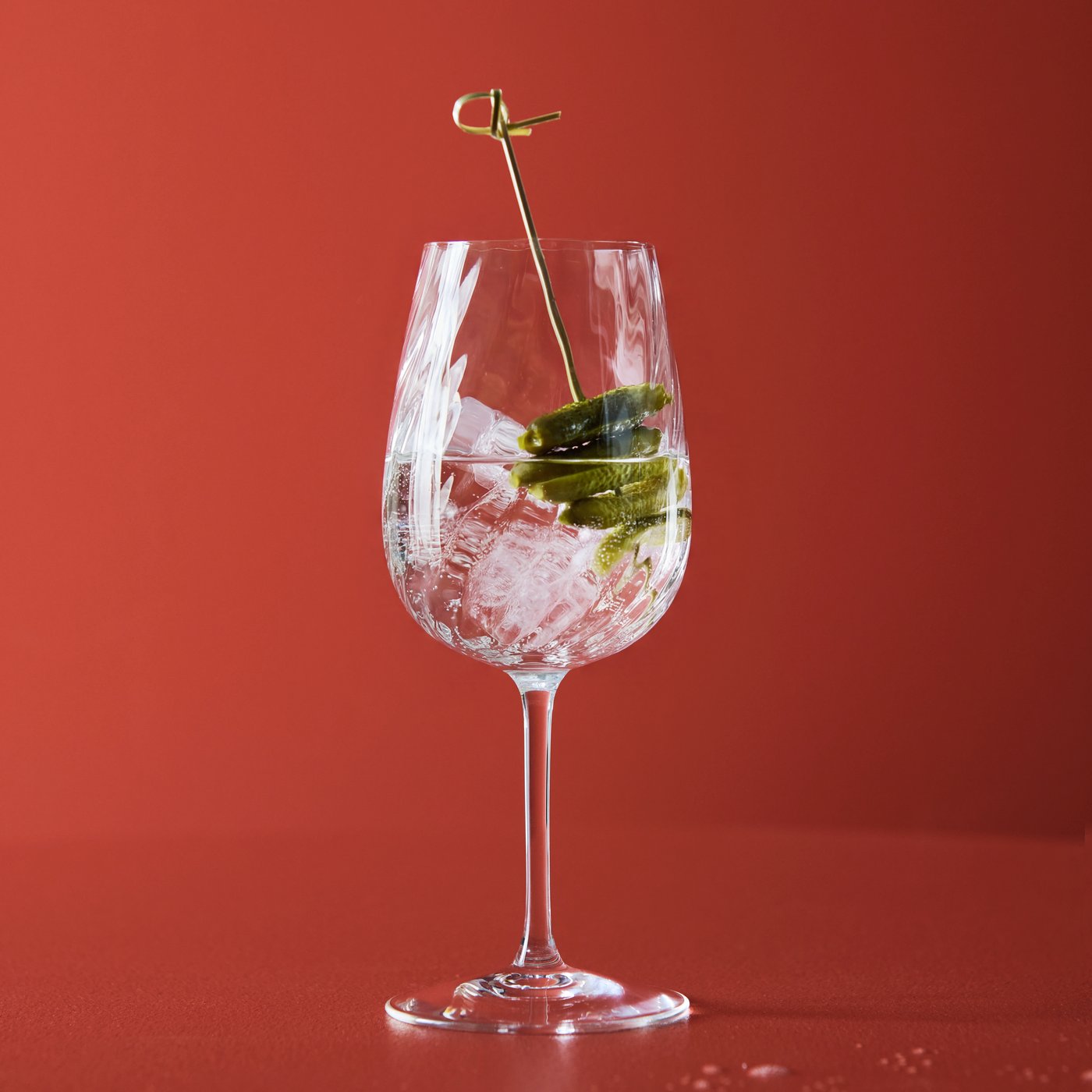 A clear FJÄLLUGGLA glass with an iced sparkling drink and pickled cucumber in it stands against a red background.