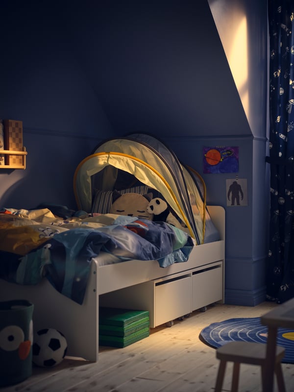 A white SLÄKT bed in a dim blue bedroom covered in blue AFTONSPARV bed linen and with an ELDFLUGA bed tent on top.