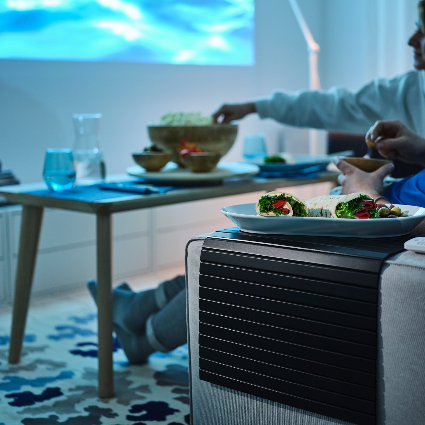 Home cinema scene with food on an IKEA RÖDEBY sofa arm tray, snacks on a low table and a film projected on the wall.