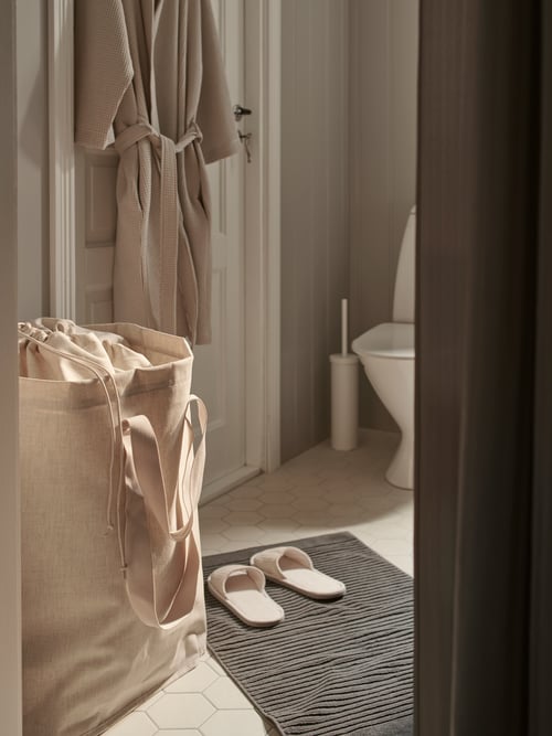 A full PURRPINGLA laundry bag by a bathroom door with a bath robe hanging on the back. A pair of slippers are on the floor.