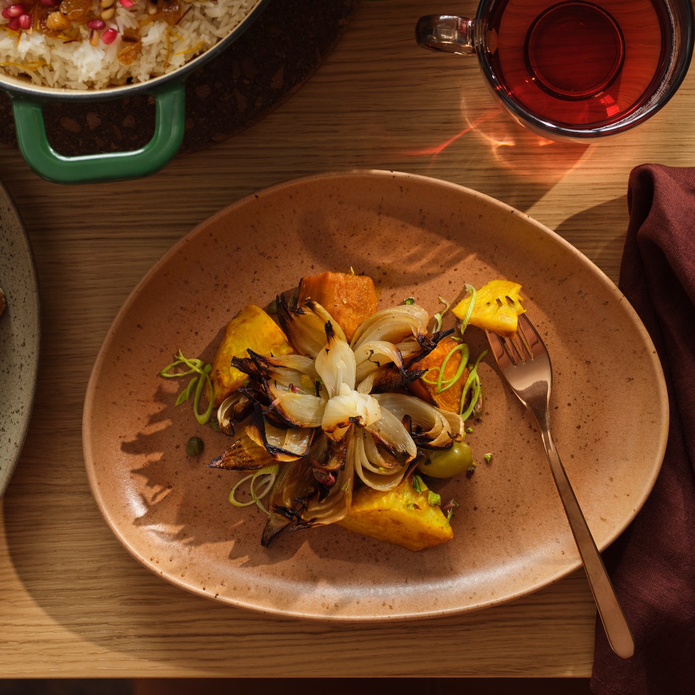 A GRÅRÄV serving plate in medium brown with roasted vegetables stands on a wooden dining table next to a glass of tea.