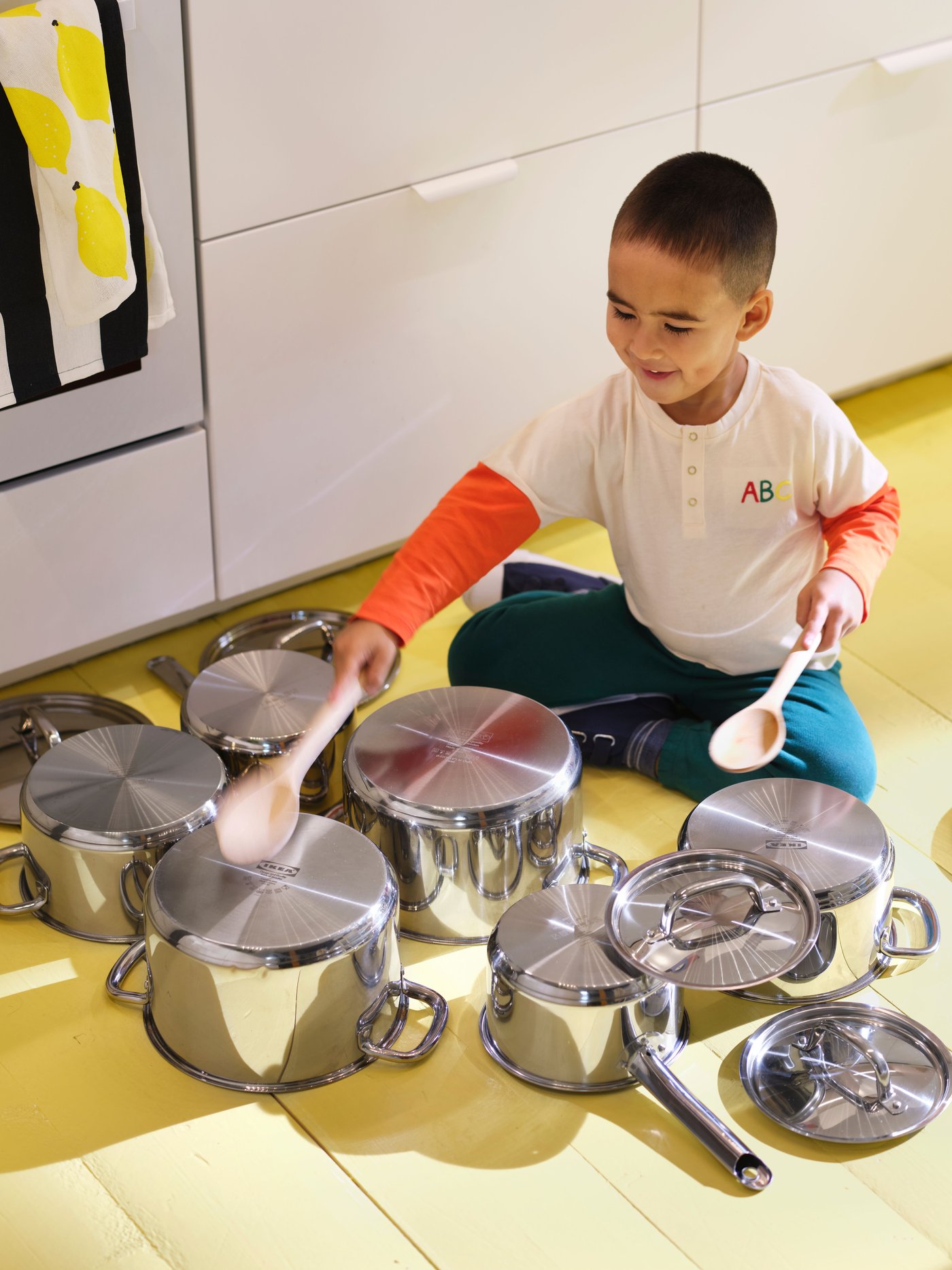 A child is sitting on a yellow kitchen floor, drumming with wooden spoons on IKEA 365+ cookware spread out on the floor.