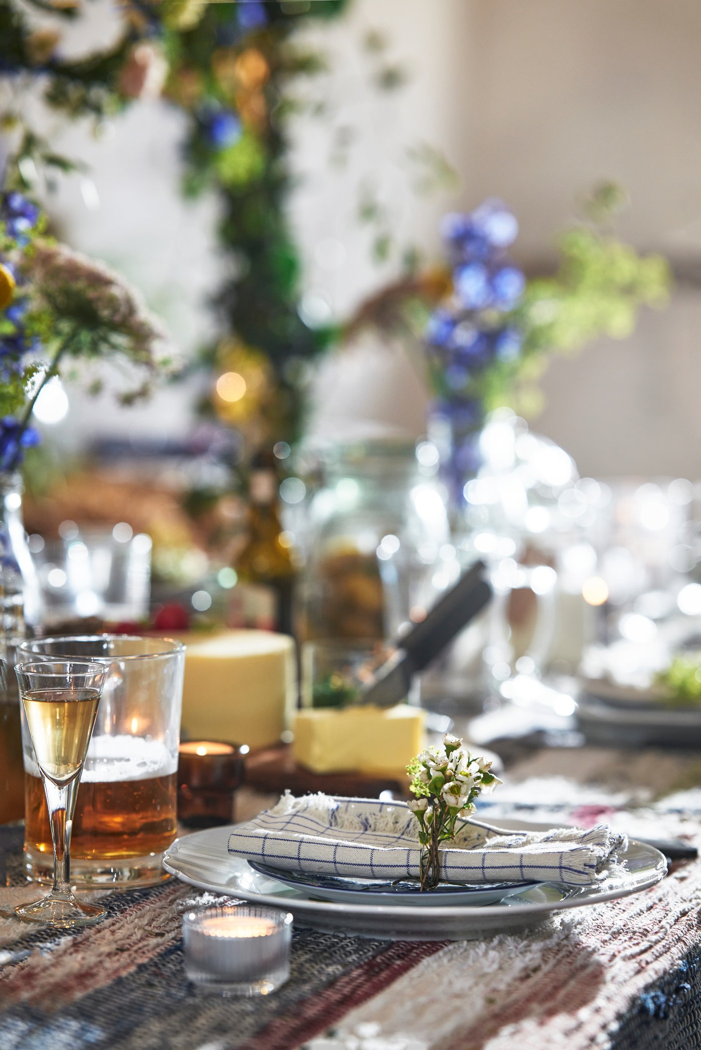 Ett rustikt dukat bord med tallrik, servett och bestick i förgrunden; runtom står glas med dryck och små ljus, och i bakgrunden syns gröna växtarrangemang och vilda blommor i ett ljust, lantligt rum.