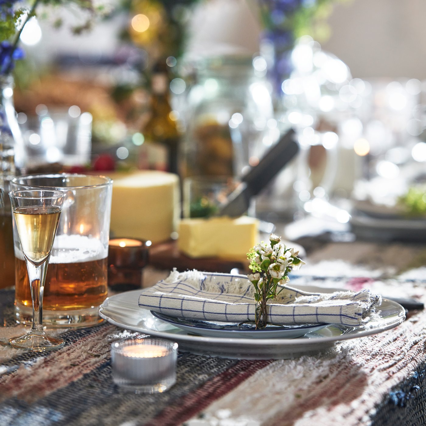 Ett rustikt dukat bord med tallrik, servett och bestick i förgrunden; runtom står glas med dryck och små ljus, och i bakgrunden syns gröna växtarrangemang och vilda blommor i ett ljust, lantligt rum.
