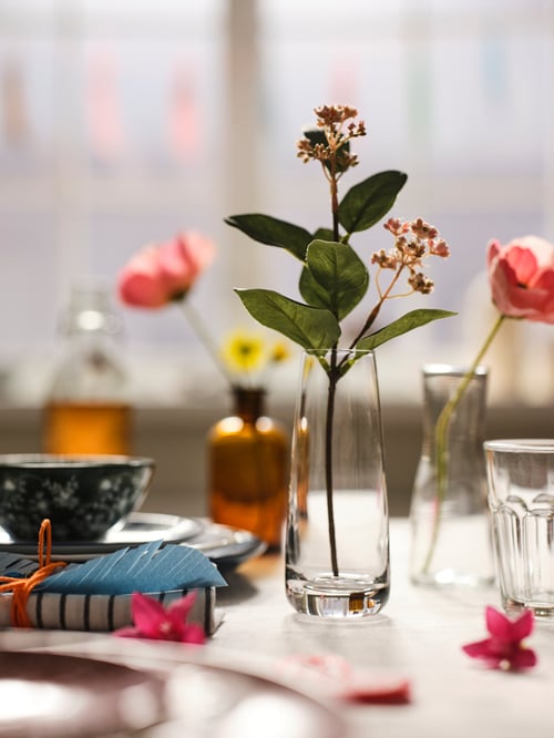 Esszimmer mit verschiedenen SMYCKA Kunstblumen in unterschiedlichen Vasen, u. a. der BERÄKNA und der TIDVATTEN Vase. Drumherum sind z. B. die ENTUSIASM Schüsseln und POKAL Gläser zu sehen.