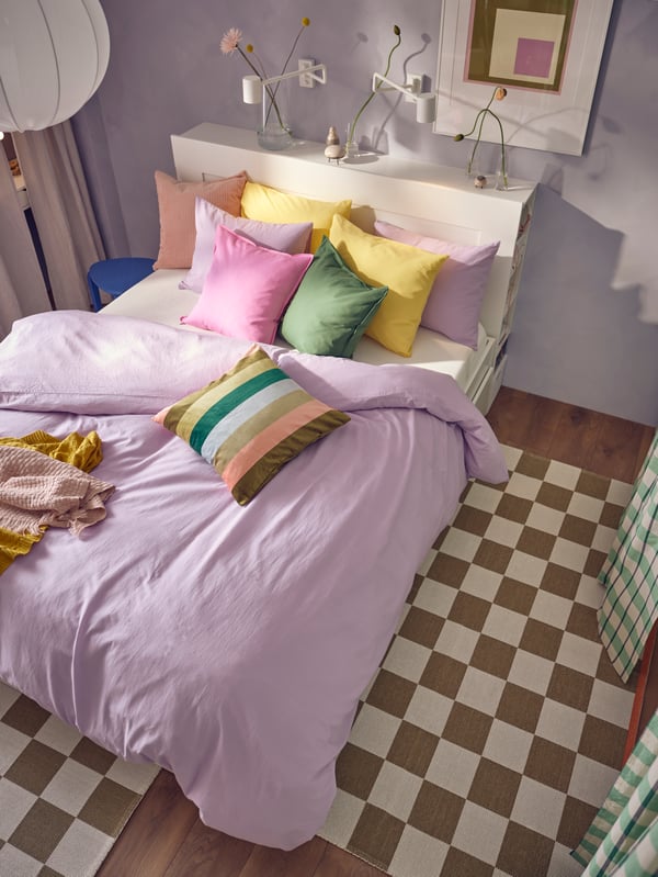 A BRIMNES bed in a lilac room covered in lilac NATTSVÄRMARE bed linen. A green GLANSHAGTORN curtain covers clothing storage.