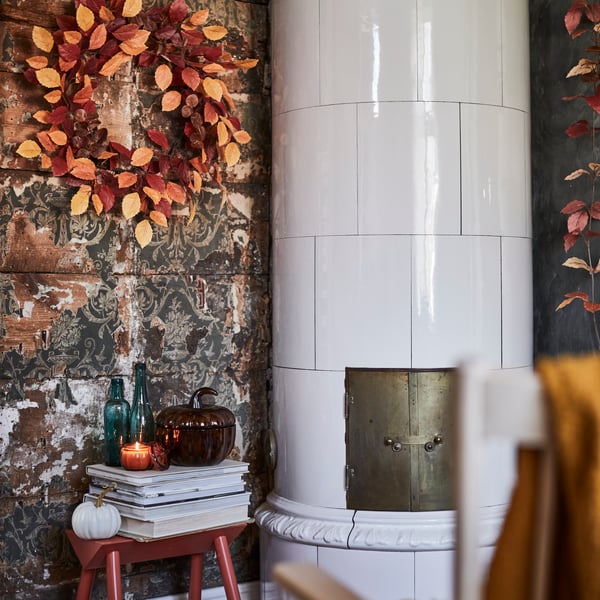 A HÖSTAGILLE artificial wreath in mixed colours adorns a rustic wall, set amidst decorative items, some books and a candle.