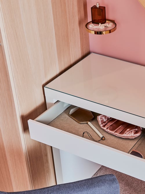 Bedroom with white dressing table with drawer, hairbrush, personal belongings, bottle of perfume placed on glass wall stand.