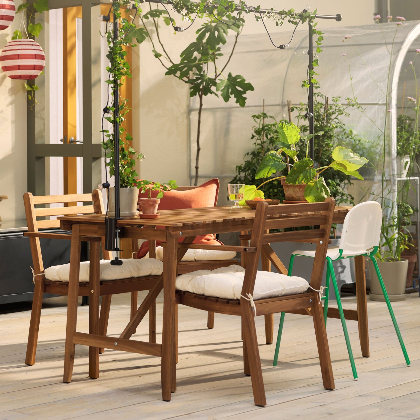 Na jasnym tarasie stoją ciemnobrązowy stół ogrodowy ASKHOLMEN, krzesła ogrodowe z podłokietnikami oraz dużo roślin.