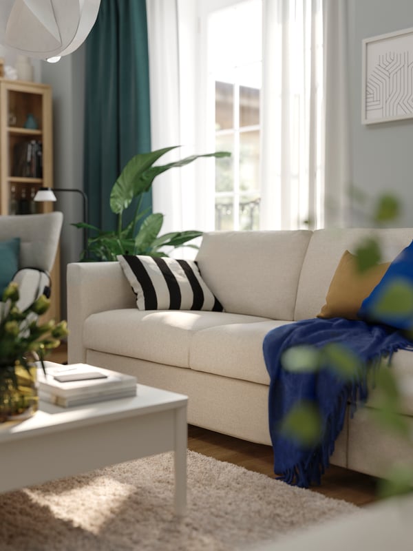 A blue KLIPPOXEL throw and some cushions lie on a beige FINNALA sofa-bed near a window in a sunny living room.