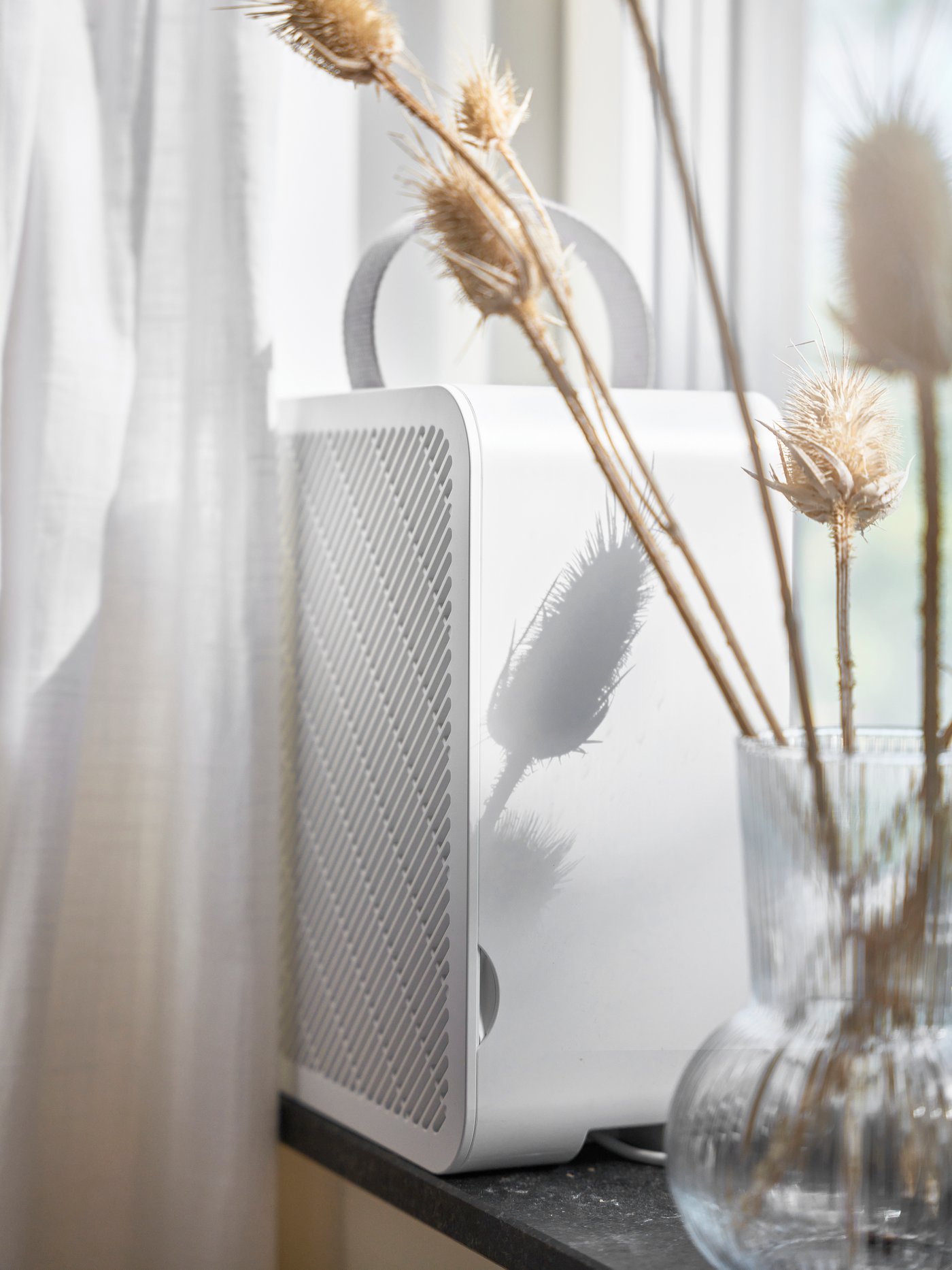 White UPPÅTVIND  air purifier with handle stands on a windowsill next to a glass vase of dried plants. Sunlight casts shadows on the appliance and the curtain.
