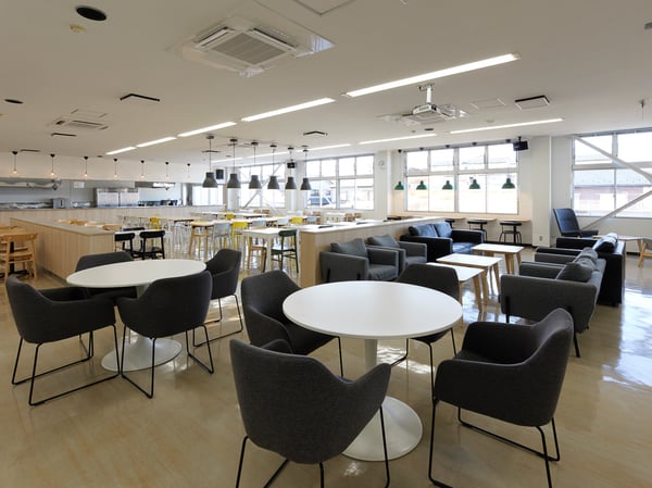 丸テーブルとグレーのチェアを配置したカフェ風の休憩スペース。奥にカウンターテーブルとペンダントライトが並ぶ明るいエリア。