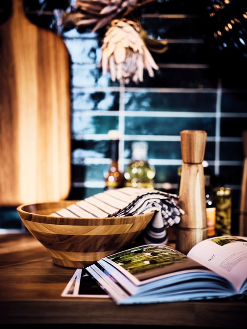 A bamboo GRÖNSAKER serving bowl, INTRESSANT spice mill and open cookbook on a wooden worktop.