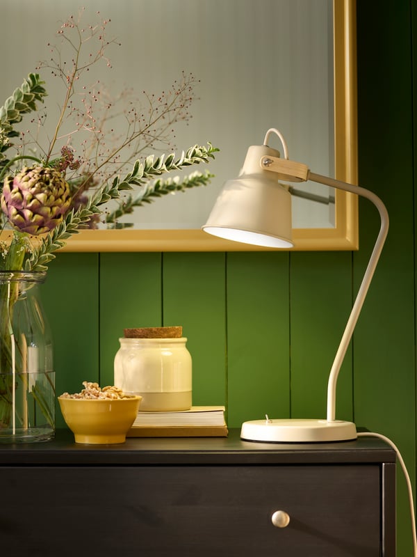 Uma candeeiro de secretária SJÖMAKT bege ilumina uma parede verde, ao lado de um vaso com flores e objetos decorativos.