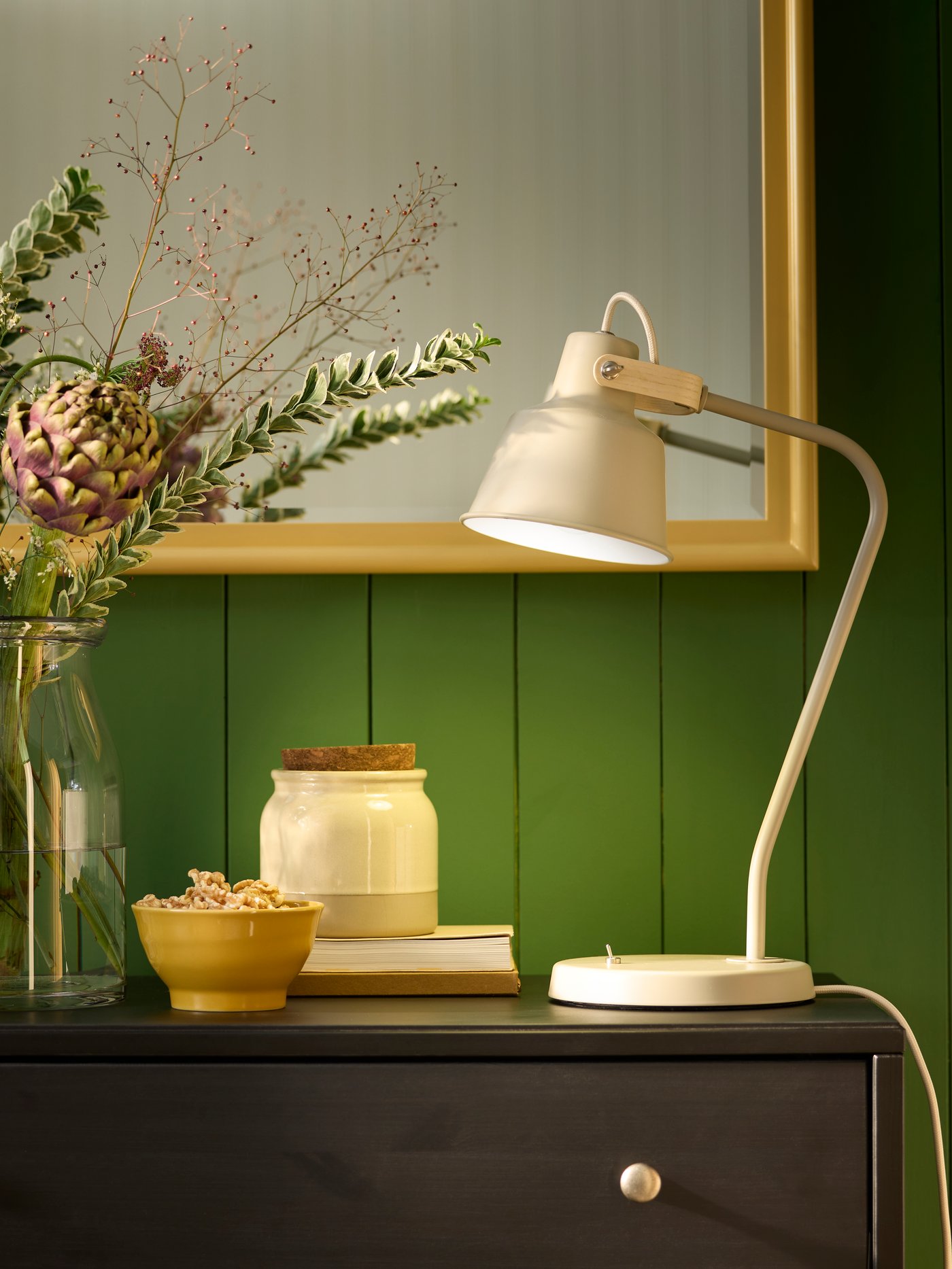 Uma candeeiro de secretária SJÖMAKT bege ilumina uma parede verde, ao lado de um vaso com flores e objetos decorativos.