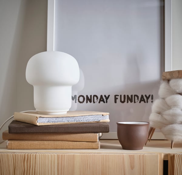 Nahaufnahme eines Bücherstapels, auf dem eine kleine Tischlampe steht, daneben eine Tasse und dahinter ein Bild im Rahmen mit dem Text "Monday Funday".