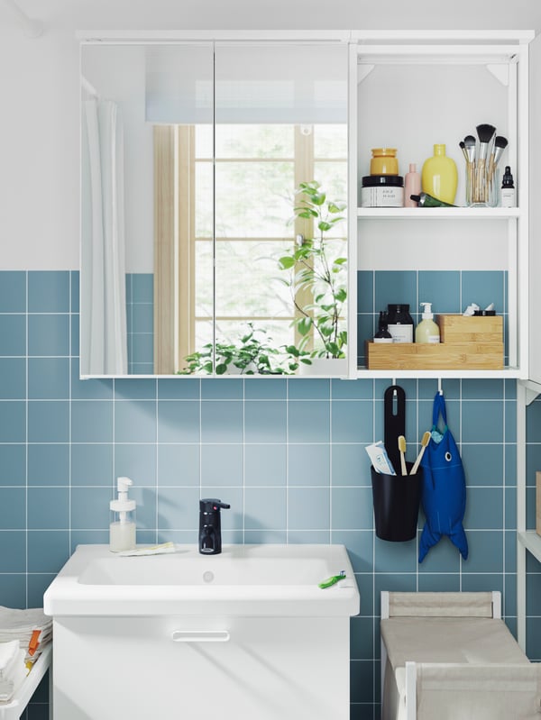 A bathroom has a white ENHET base cabinet, a wall cabinet with mirror doors and a frame with shelves for care products.