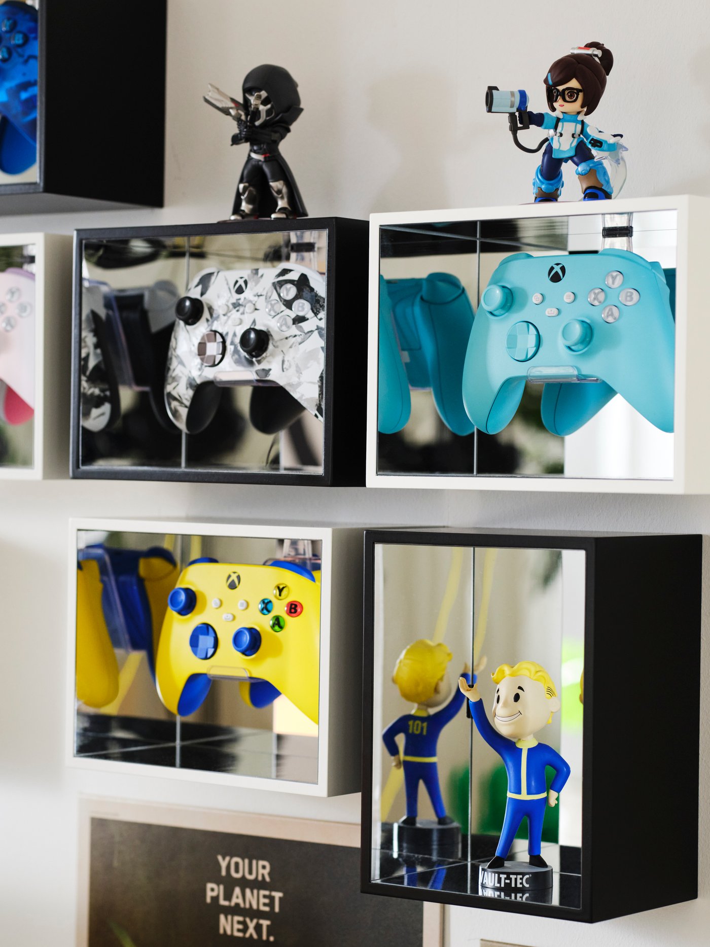 Zes YXSTABY displayboxen voor gamingcontrollers hangen aan een witte muur, met controllers in verschillende kleuren en collectibles erin.