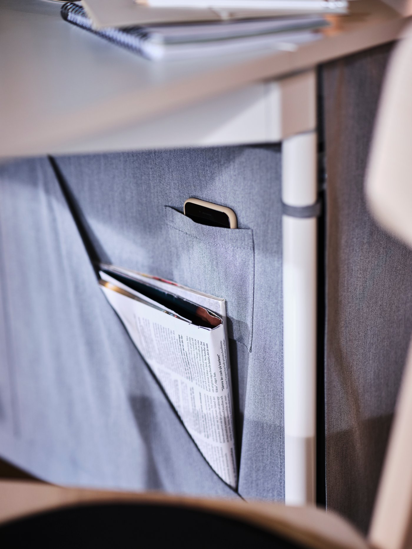 The under-table storage of a white/light grey KALLHÄLL gateleg table, holding a newspaper and mobile phone.