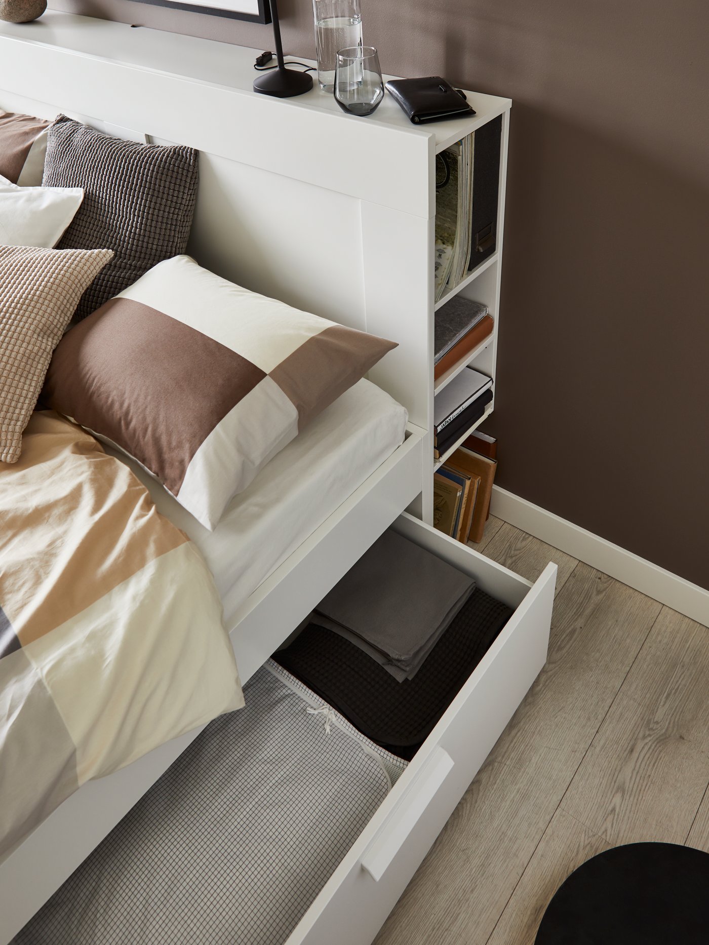 収納付きヘッドボードと引き出しを備えた白いベッド。ブラウン系の寝具と木目調の床が温かみを演出。