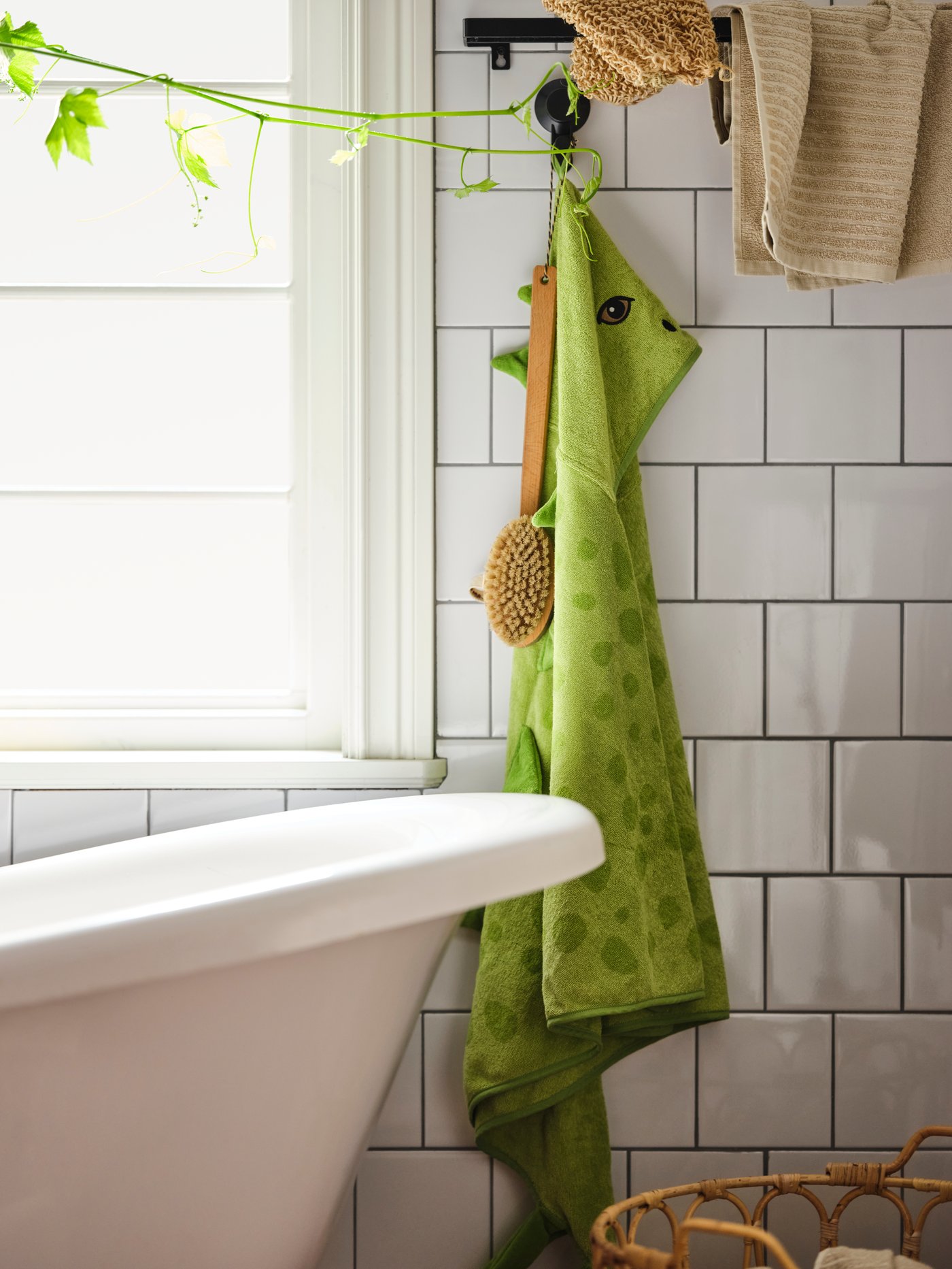 A green JÄTTELIK stegosaurus towel with hood hangs from a hook next to a bath in a white-tiled bathroom.