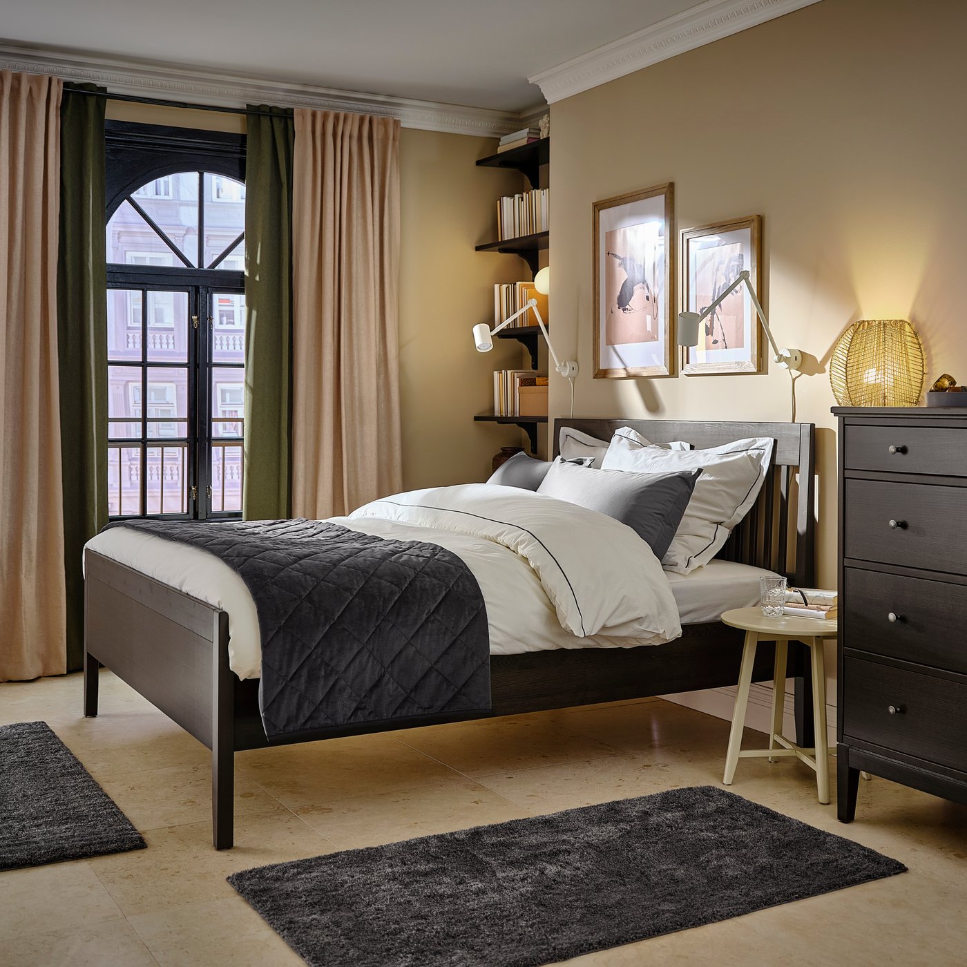 Un dormitori en tons marrons amb mobles negre fosc, que inclou una estructura de llit, catifes, mobles de paret i decoració, així com un conjunt de taules niu KRAGSTA en beix clar.