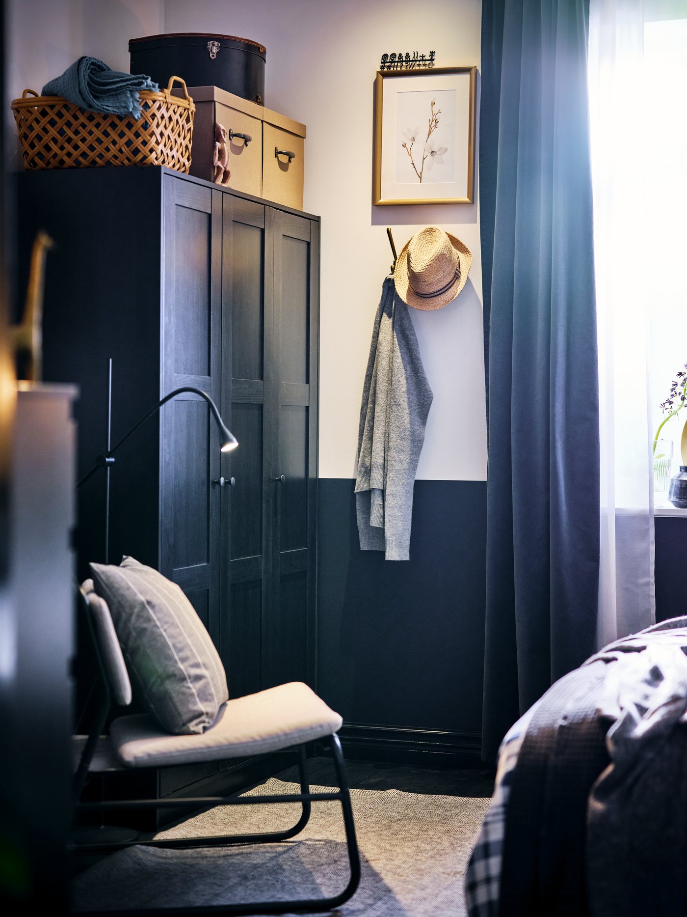 A corner of a bedroom with a black-brown RAKKESTAD wardrobe and a light-grey LINNEBÄCK easy chair standing next to it.