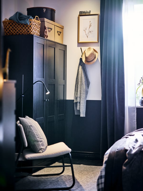 A corner of a bedroom with a black-brown RAKKESTAD wardrobe and a light-grey LINNEBÄCK easy chair standing next to it.