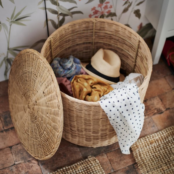 A TOLKNING pouffe with storage on a wooden floor with the lid off and leaning against it revealing hats and scarves inside.