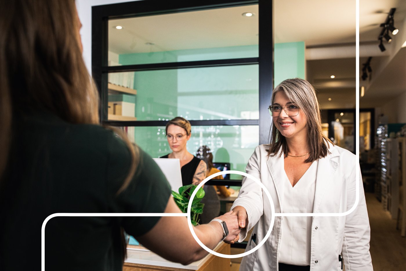 Två personer skakar hand i ett kontorsmiljö, med en tredje person sittande vid ett skrivbord i bakgrunden.