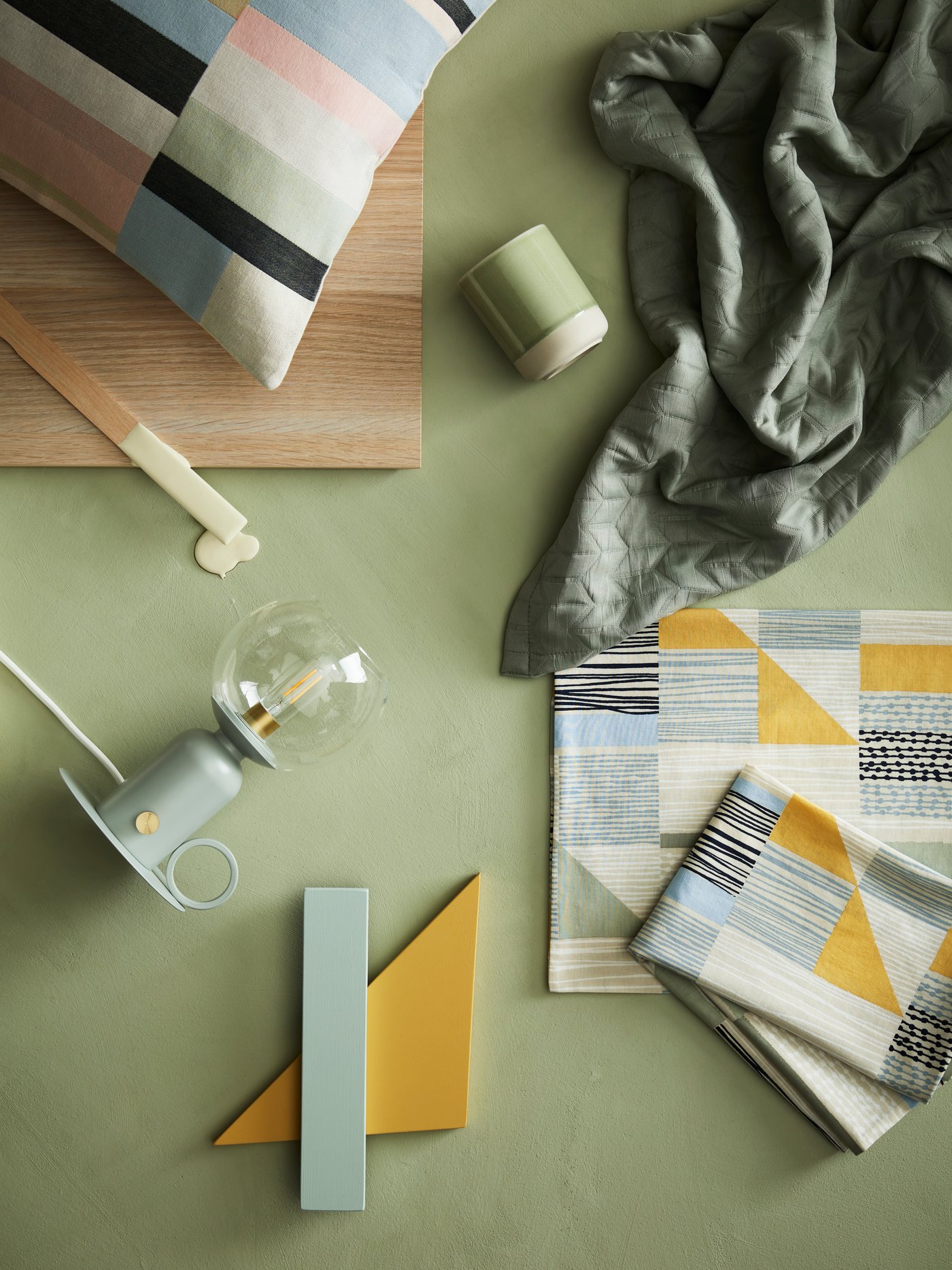 An artistic display with a printed cushion, candle holder, bedside lamp and folded pillowcases on a light-green surface.