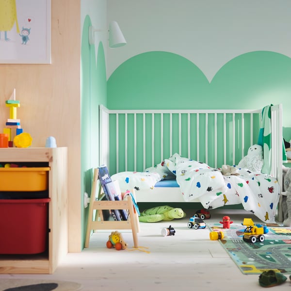 A toddler’s room with green clouds painted on the wall, GULLIVER cot in back, VALLABY rug and lots of toy cars on the floor.
