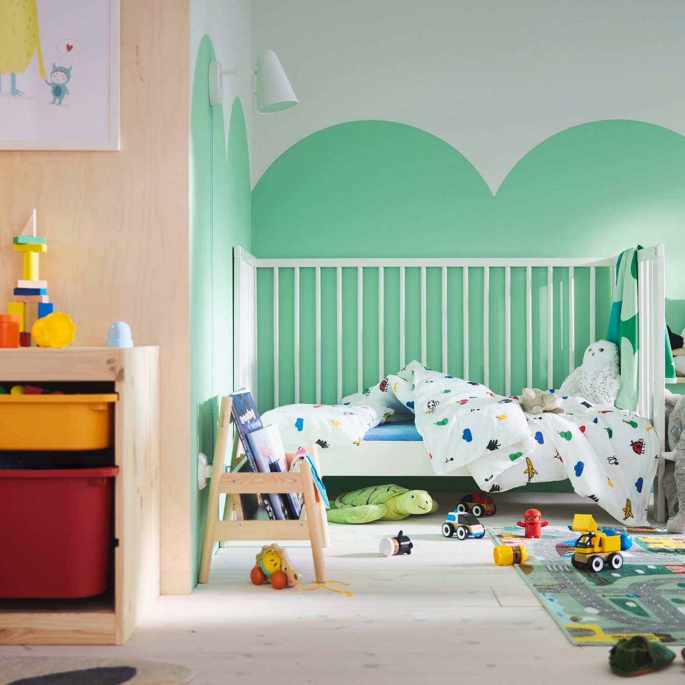 A toddler’s room with green clouds painted on the wall, GULLIVER cot in back, VALLABY rug and lots of toy cars on the floor.