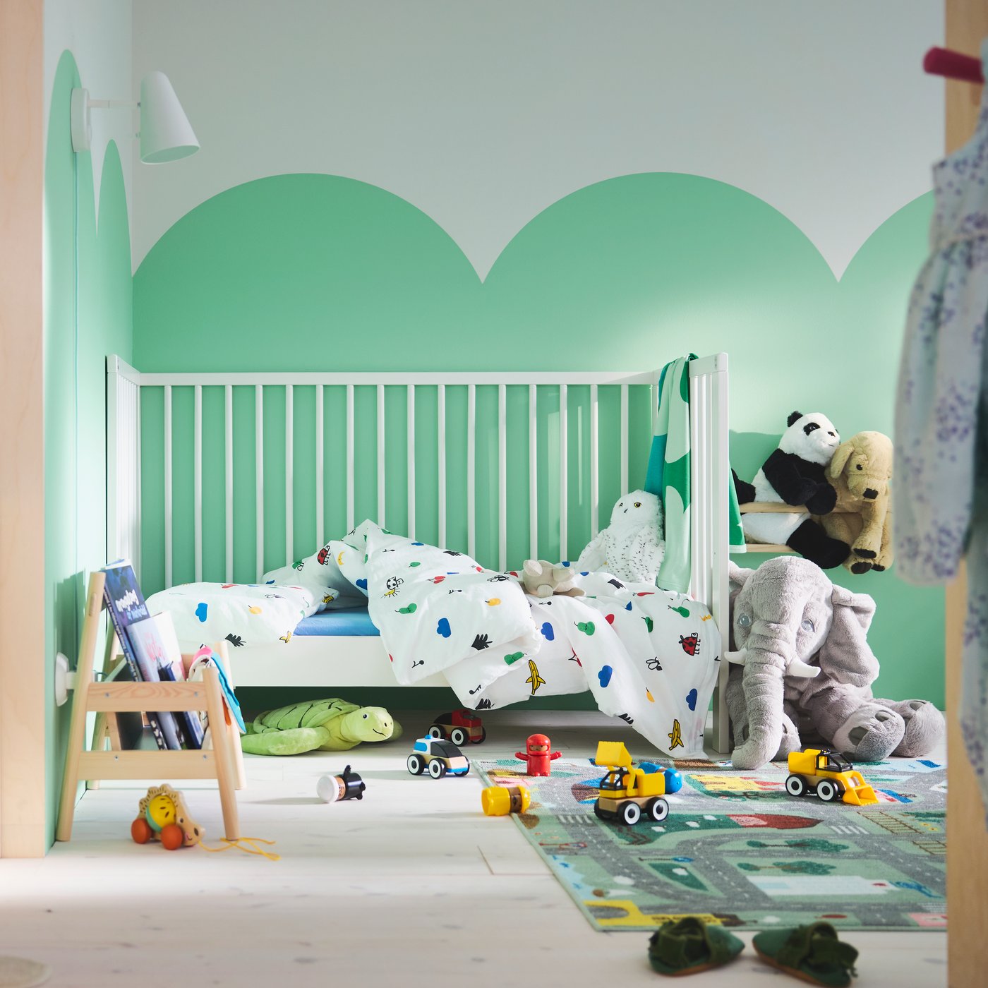 ห้องของเด็กวัยหัดเดินที่มีรูปก้อนเมฆสีเขียววาดบนผนัง, เตียงเด็กอ่อน GULLIVER/กุลลิแวร์ ที่ด้านหลัง, พรม VALLABY/วัลลาบี และรถของเล่นหลายคันบนพื้น