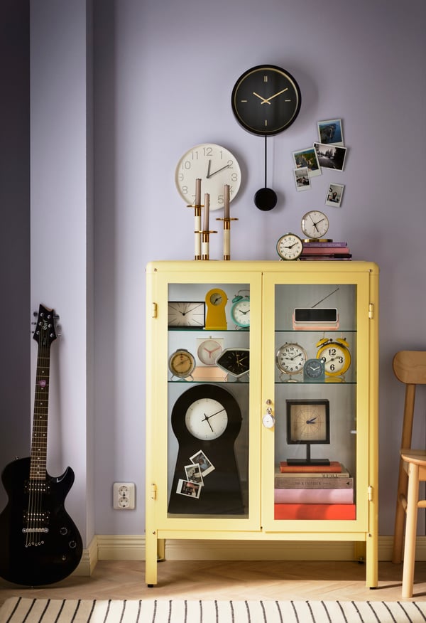 A yellow cabinet with glass doors with varius alarms and clocks in the living room.
