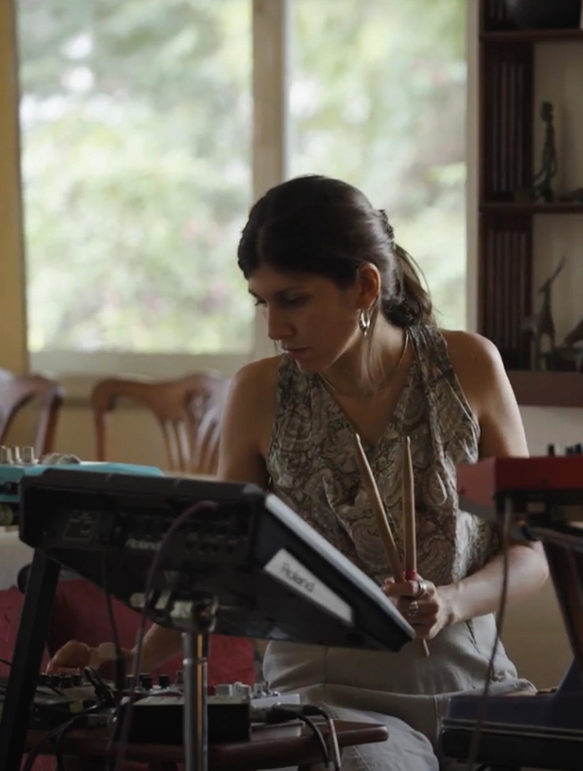 Žena sedi pred ekranom uređaja i drži palice za bubnjeve.