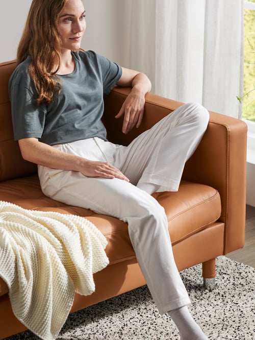 A woman in a living room sits on a three-seat sofa next to a white throw with one foot on a rectangular high-pile rug.