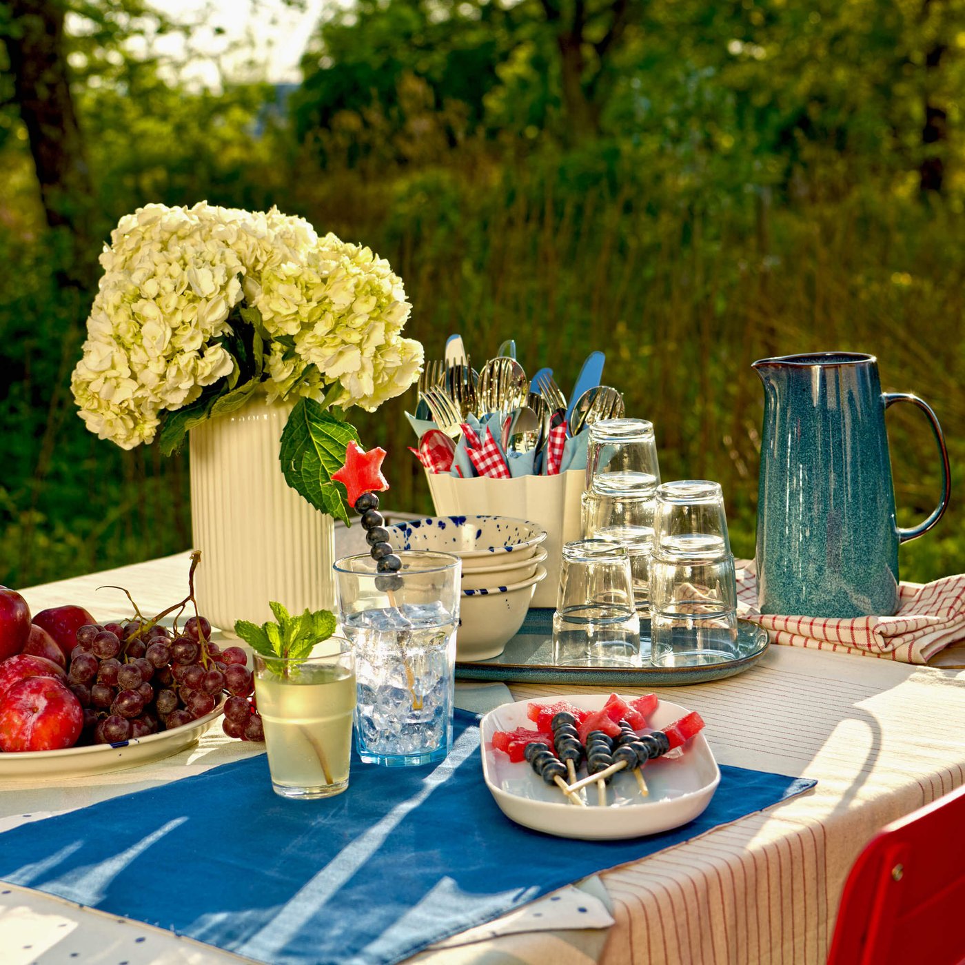 Outdoor picnic table with flowers, fruit, drinks and skewered berries in a garden setting.