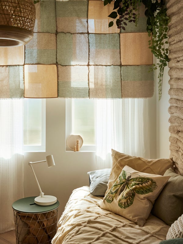 Boho deco style daybed with throw and scatter cushions in front of white-curtained window with patchwork wall-hanging and desk lamp on a storage table.