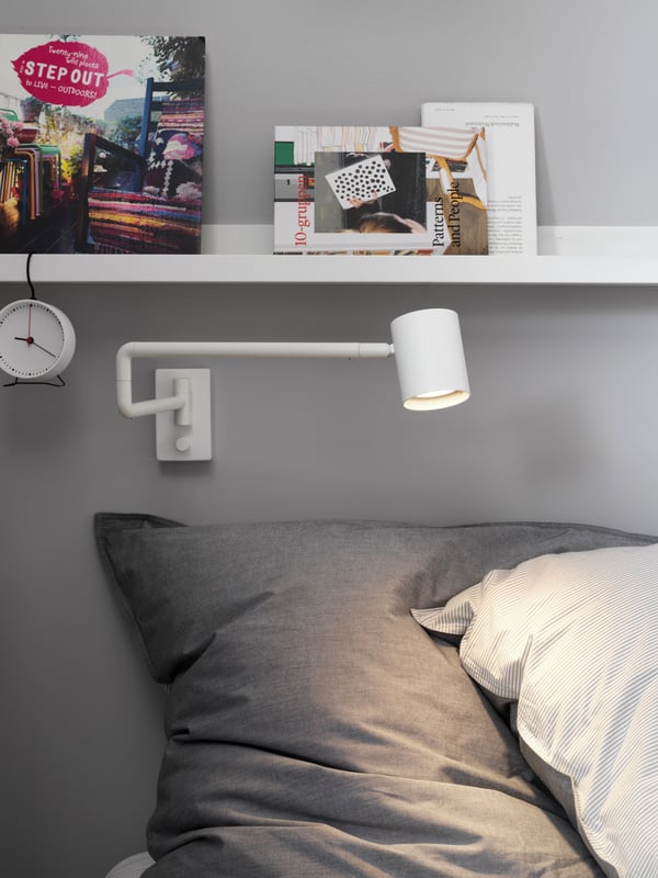 La cabecara de una cama con una lámpara de pared apuntando a la almohada. Sobre la lámpara hay una repisa de pared con unos vinilos y unos cuadros pequeños.