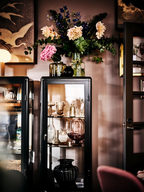 A FABRIKÖR glass-door cabinet filled with glassware, with vases of fresh flowers on top and a framed print above.