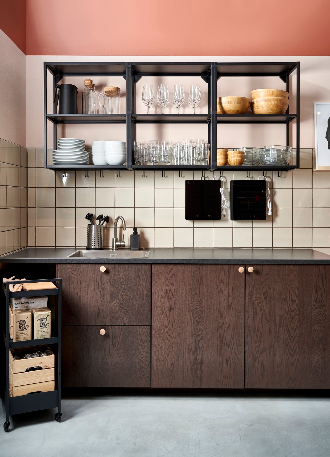A kitchen with wooden panel doors and a shelving unit above seen from the front. There is a black trolley to the left with boxes.