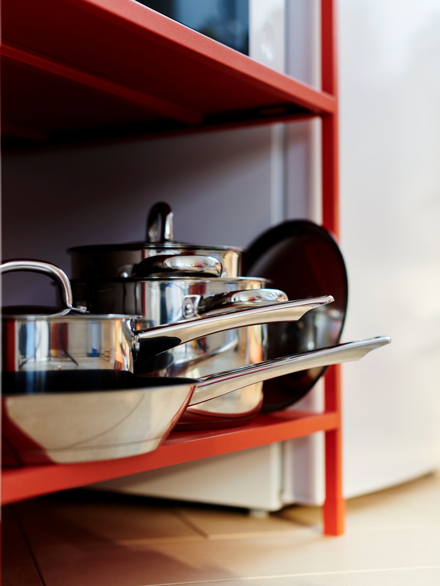 Dua periuk, satu periuk bertangkai dan satu kuali leper dengan penutup keluli tahan karat disimpan di para bawah unit rak berwarna merah.