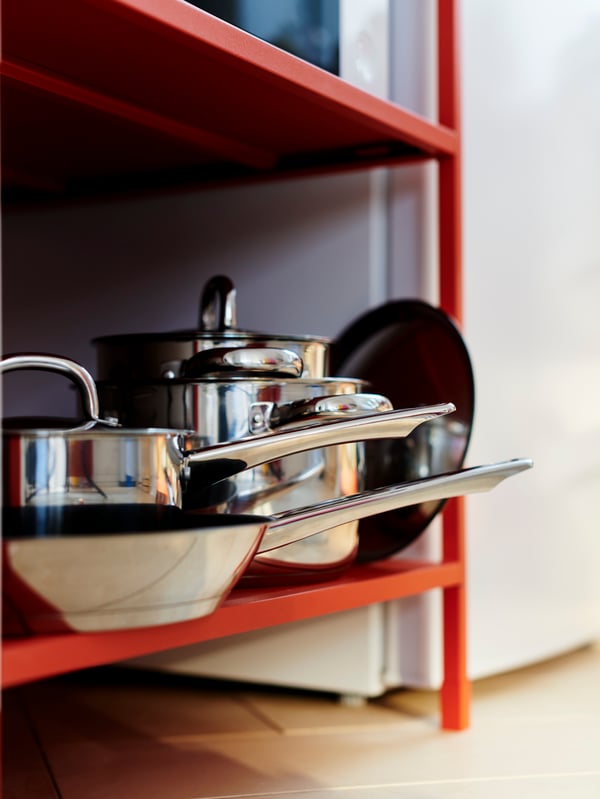 Two pots, one saucepan and one frying pan with lids in stainless steel are stored on the bottom shelf of a red shelving unit.