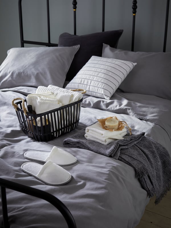 Sänky, jossa on harmaat petivaatteet ja jonka päällä on valkoisia pyyhkeitä ja aamutossut