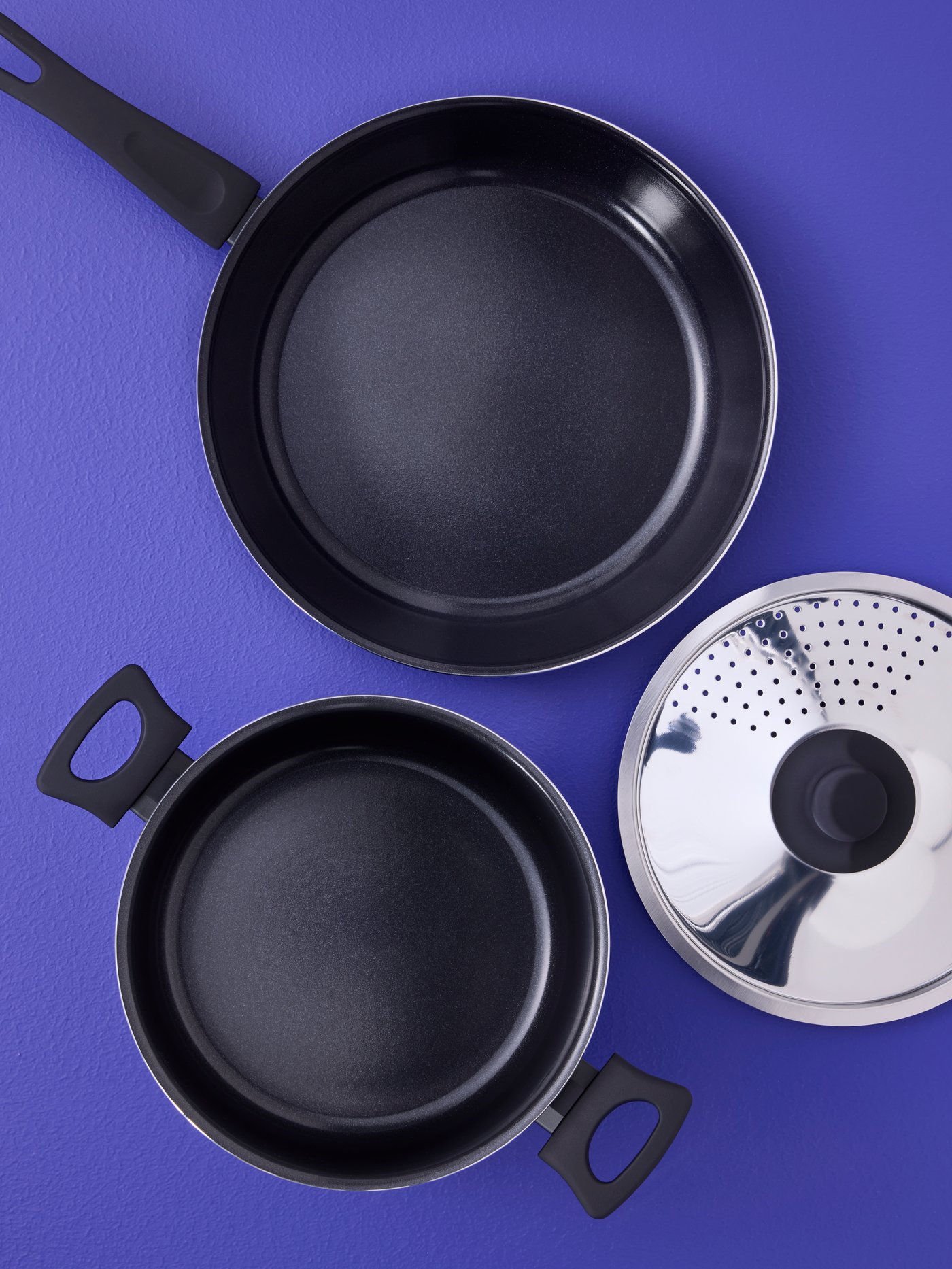 A black HEMGLAGAD pot with lid and a black IKEA 365+ frying pan, both with non-stick coating, are placed on a blue surface.