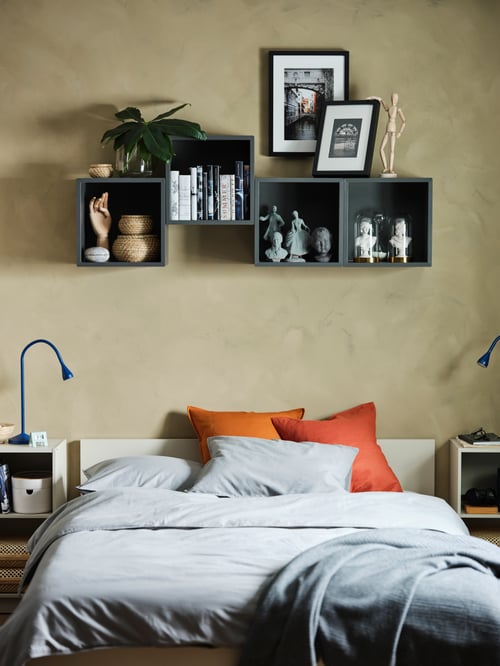 Grey, wall-mounted shelving units are above a bed frame with headboard on which is a duvet set and coloured pillows.