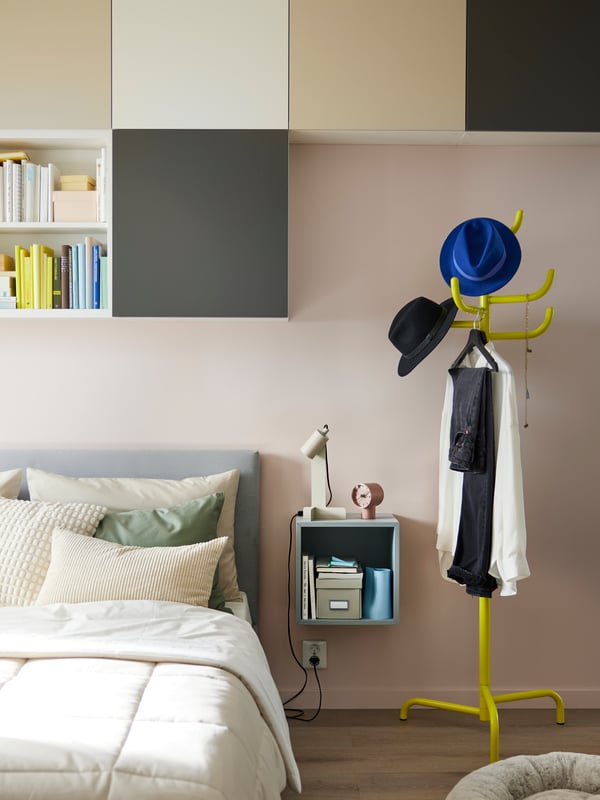 A yellow BONDSKÄRET coat stand beside a grey GLADSTAD bed covered in grey and cream textiles, with an EKET cabinet beside it.